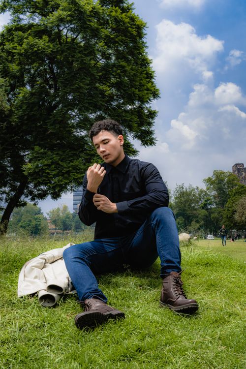Free A fashionable man in a park, adjusting his jacket, under a blue sky. Stock Photo
