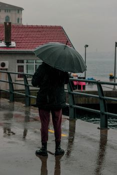 Individual stands with umbrella by a waterfront on a rainy day, capturing a serene moment.