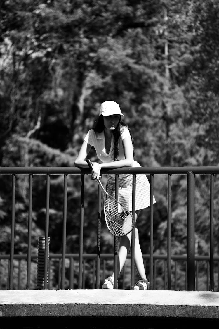 Grayscale Photo Of Woman In Tennis Outfit Leaning On Metal Railing