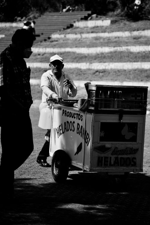 Free Black and white image of a vendor pushing a vintage ice cream cart in an urban area. Stock Photo