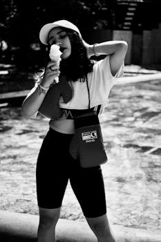 A woman in a baseball cap savors ice cream on a sunny day, captured in black and white.