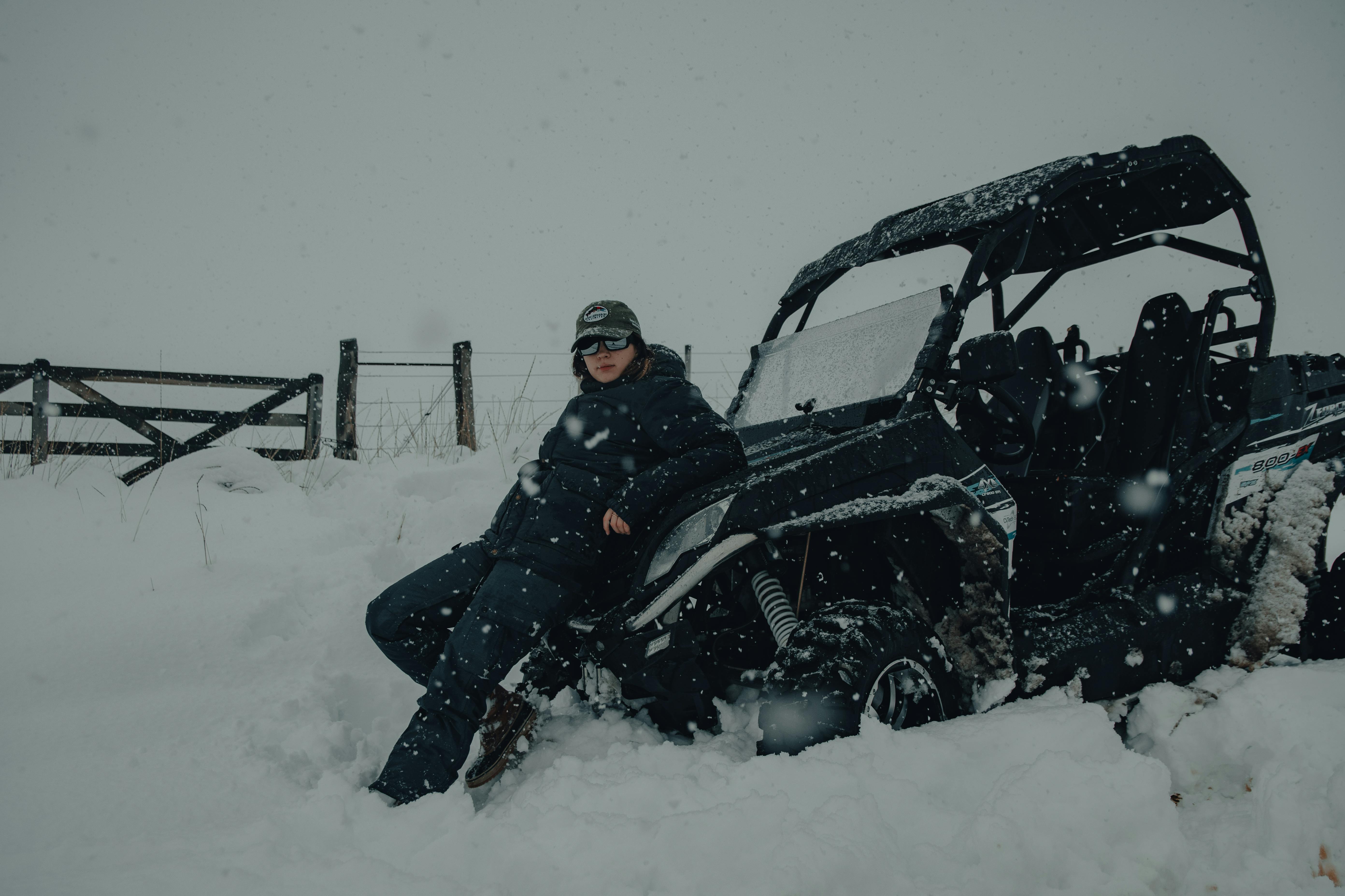 Woman in warm clothing leaning on an ATV in snowy winter countryside setting.