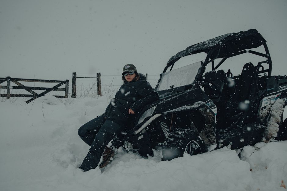 Woman in warm clothing leaning on an ATV in snowy winter countryside setting.