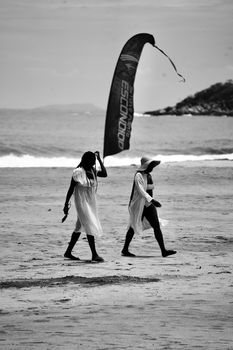 Two people walking on a sandy beach in black and white with a wind sail in the background.