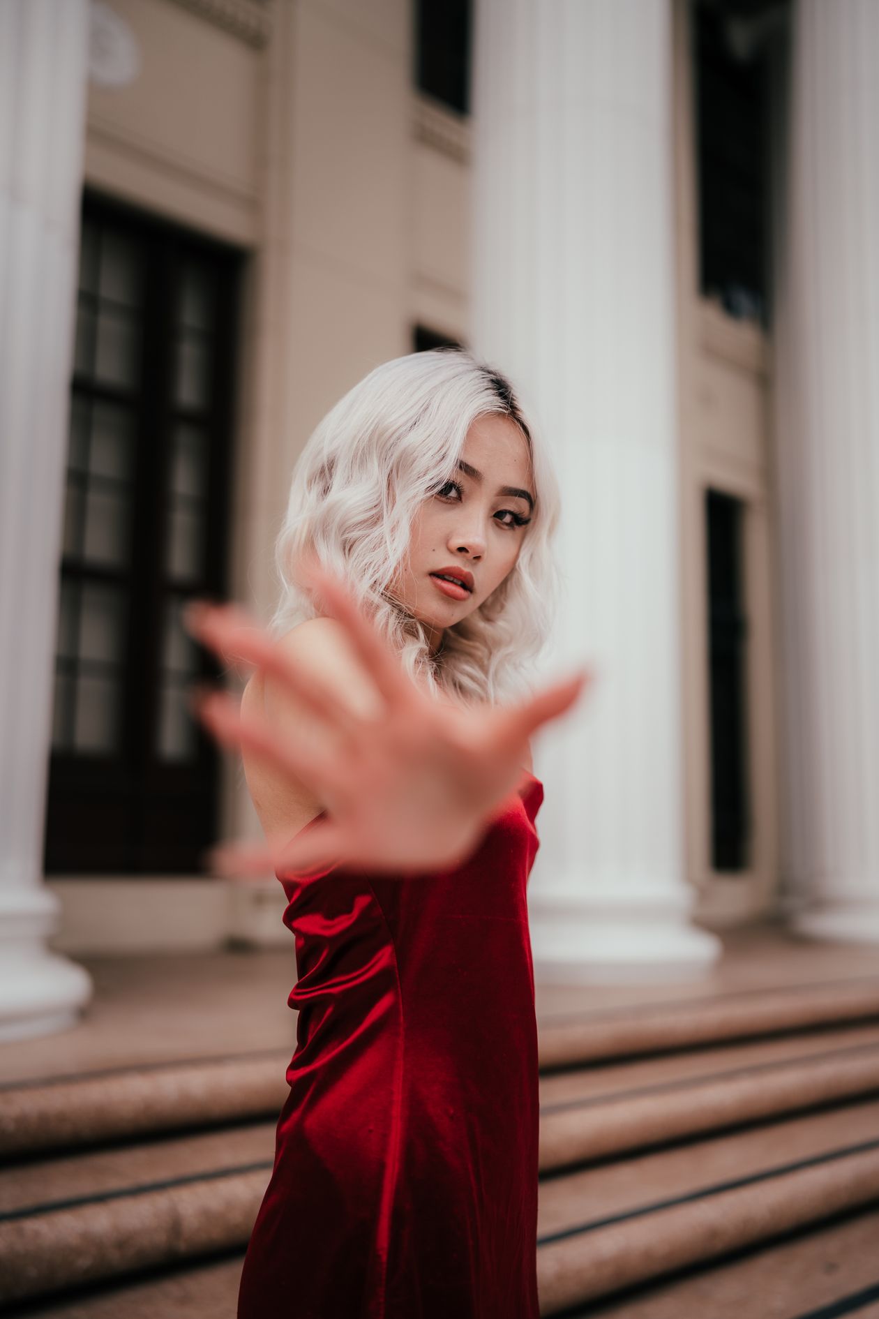 Woman in a deep red satin dress reaching toward the camera in front of stone columns