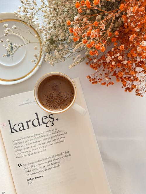 A Cup of Coffee on an Open Book Beside Dried Flower on a White Surface