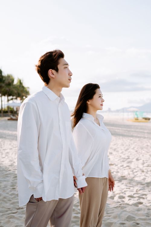 Free Asian couple enjoying a peaceful walk on a sunny beach, holding hands and looking towards the horizon. Stock Photo