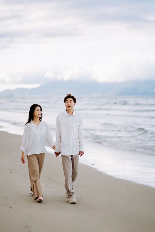 Free Asian couple walking hand in hand on a peaceful beach, enjoying a romantic day together. Stock Photo