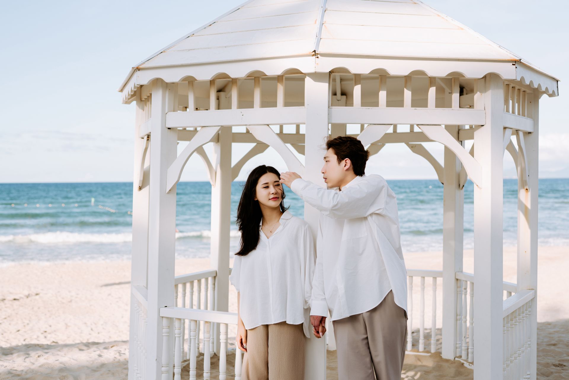 Asian couple in white shirts sharing a romantic moment under a beach gazebo by the sea.