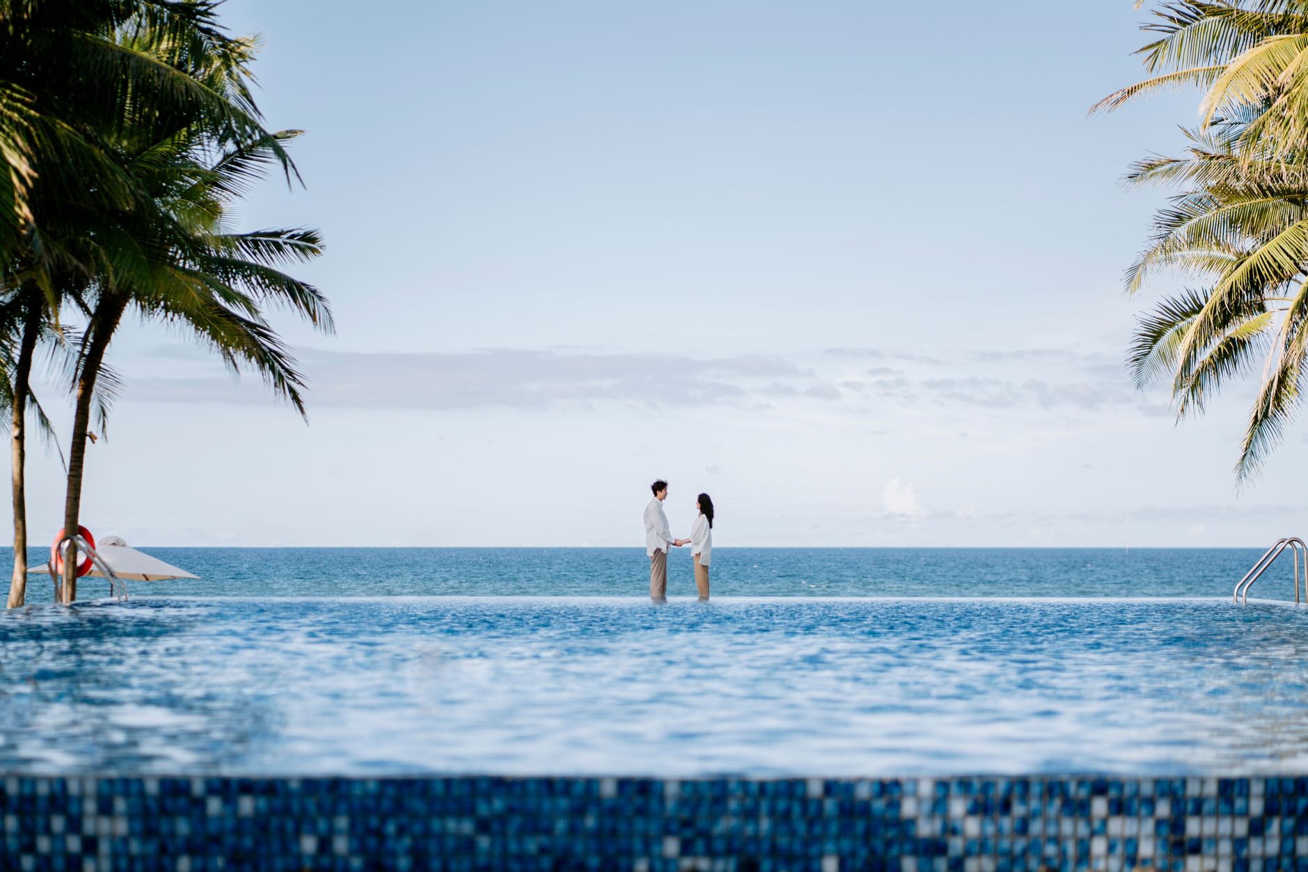 A couple stands by an infinity pool, gazing at the tropical ocean on a sunny day.