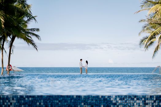 A couple stands by an infinity pool, gazing at the tropical ocean on a sunny day.