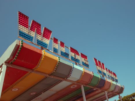 Vibrant retro amusement park sign "Wilde Maus" with colorful roof against clear blue sky.
