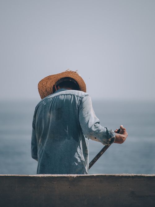 Free A dedicated worker in a denim jacket and straw hat stands by the ocean, symbolizing manual labor. Stock Photo