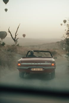 A classic red convertible travels on a dusty road with hot air balloons in the background.