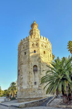The historic Torre del Oro tower in Seville, Spain under a clear blue sky, surrounded by palm trees.