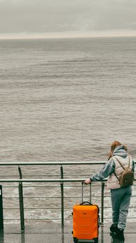 A woman in winter clothing leans on a railing with luggage, gazing at the overcast sea in Sochi, Russia.