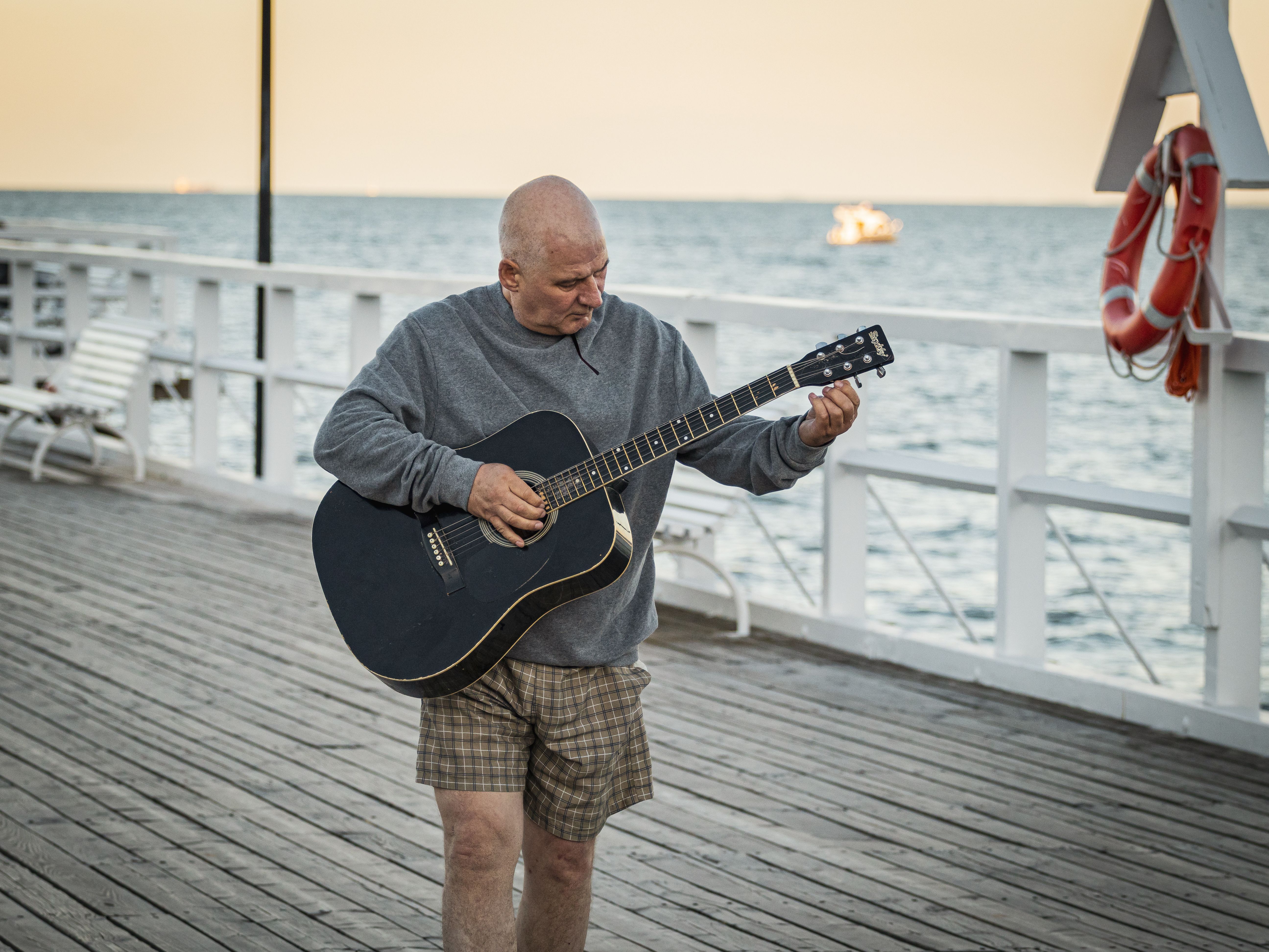A bald man plays an acoustic guitar on a wooden pier by the sea, creating a serene scene.