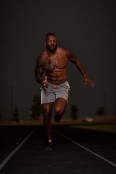 A muscular, topless man running powerfully on a track at night, showcasing strength and determination.