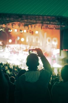 Back view of a crowd at a lively music festival with a fan recording the performance on a smartphone.