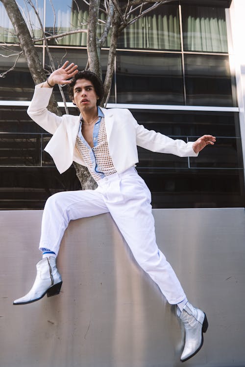 Man Wearing White Clothing Sitting on a Metal Wall and Posing in front of a Building