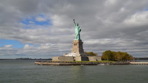 Free Iconic Statue of Liberty with New York skyline in the background under dramatic clouds. Stock Photo