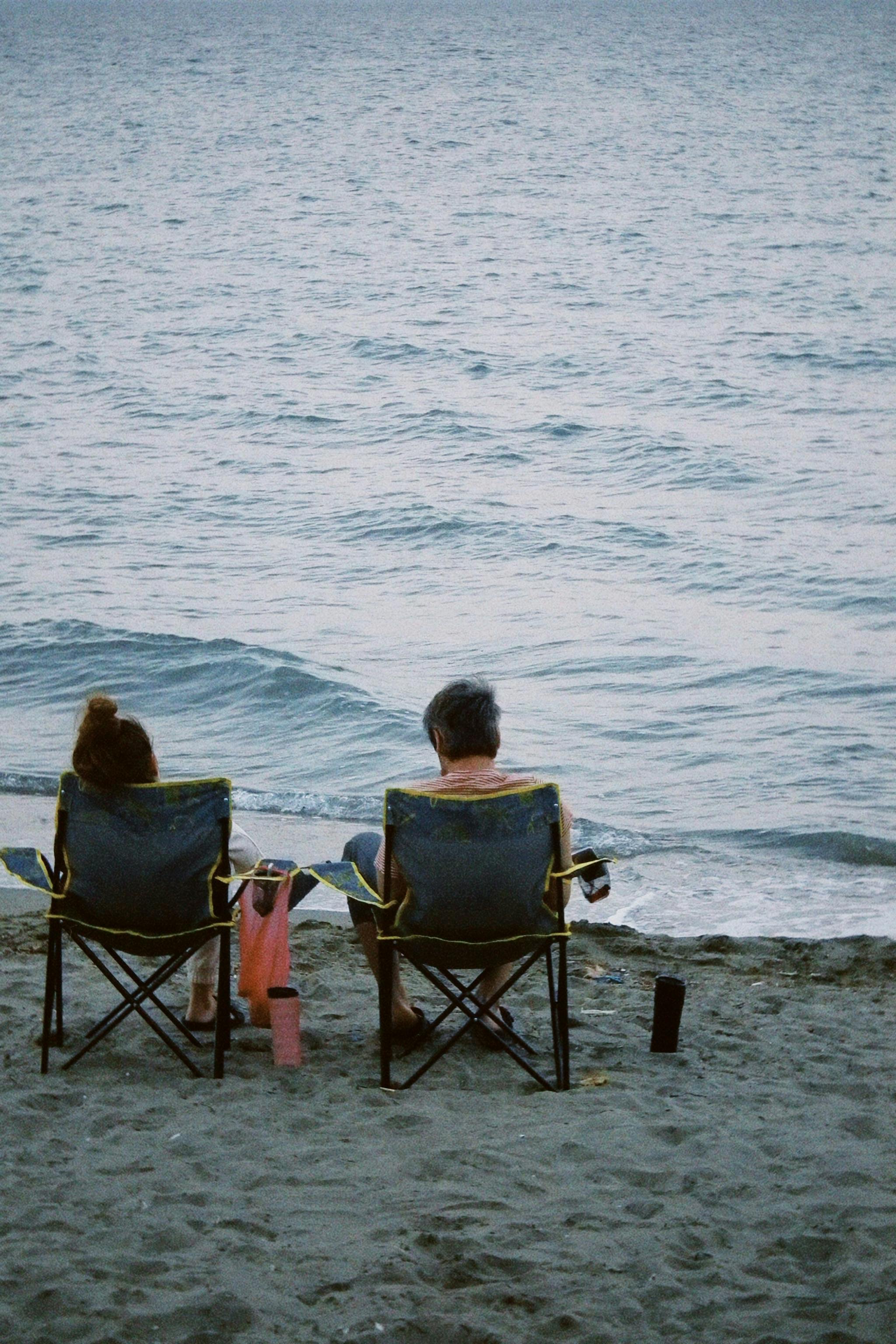 Free A couple enjoys a peaceful moment at the beach, sitting on chairs facing the sea. Stock Photo
