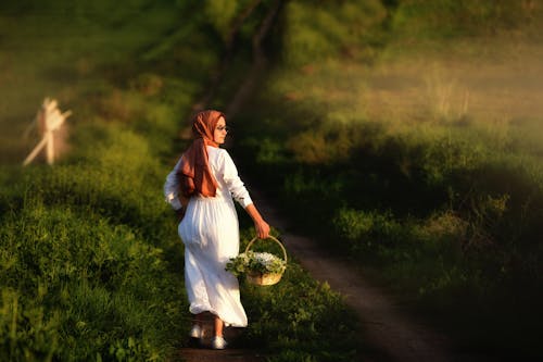 Woman in White Dress Walking on a Pathway while Carrying a Basket of Flowers