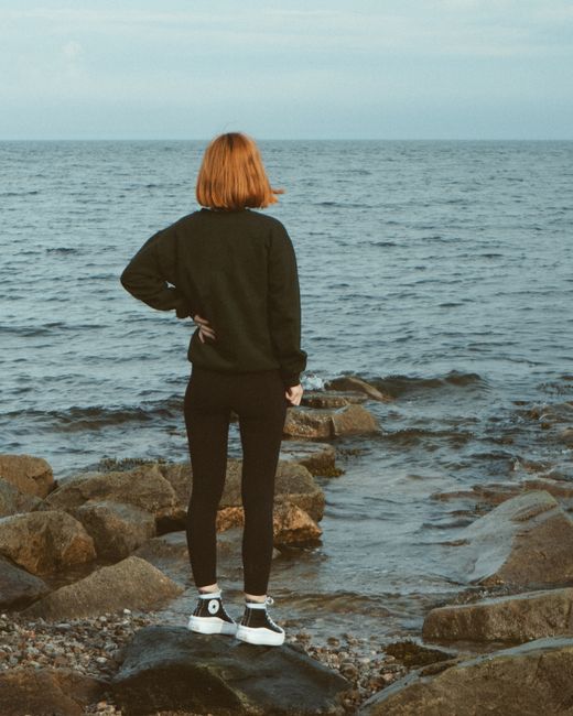 A woman stands facing the sea, hand on hip, on a rocky seashore.
