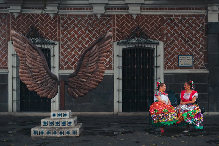 Women In Floral Dresses Sitting Near Wings Statue In Barrio Del Artista, Puebla, Mexico