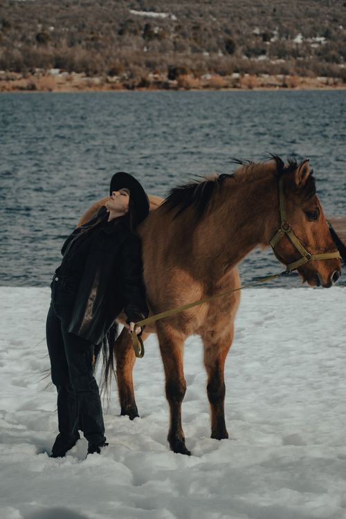 Free A woman stands beside a horse on a snowy riverside, creating a serene winter scene. Stock Photo