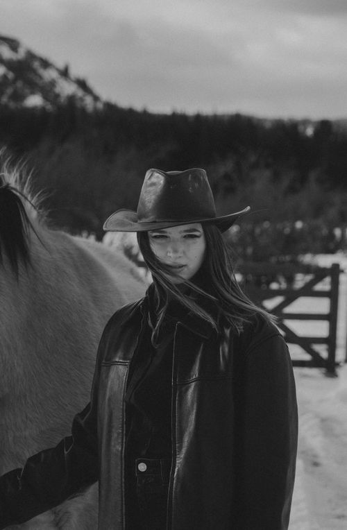 Free A striking black and white portrait of a fashionable cowgirl in a leather jacket, posing in a snowy outdoor setting. Stock Photo