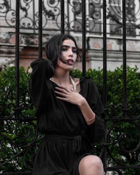 Stylish woman in a black dress and necklace posing elegantly outdoors.
