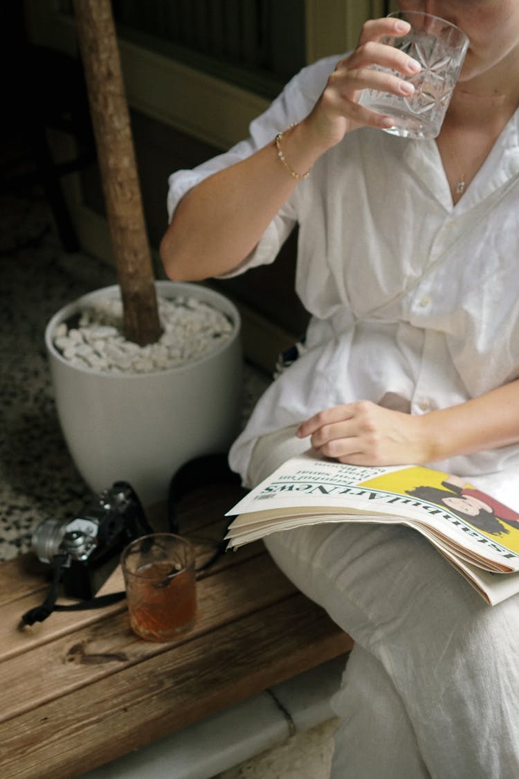 Woman Sitting, Drinking A Glass Of Water And Reading A Paper 