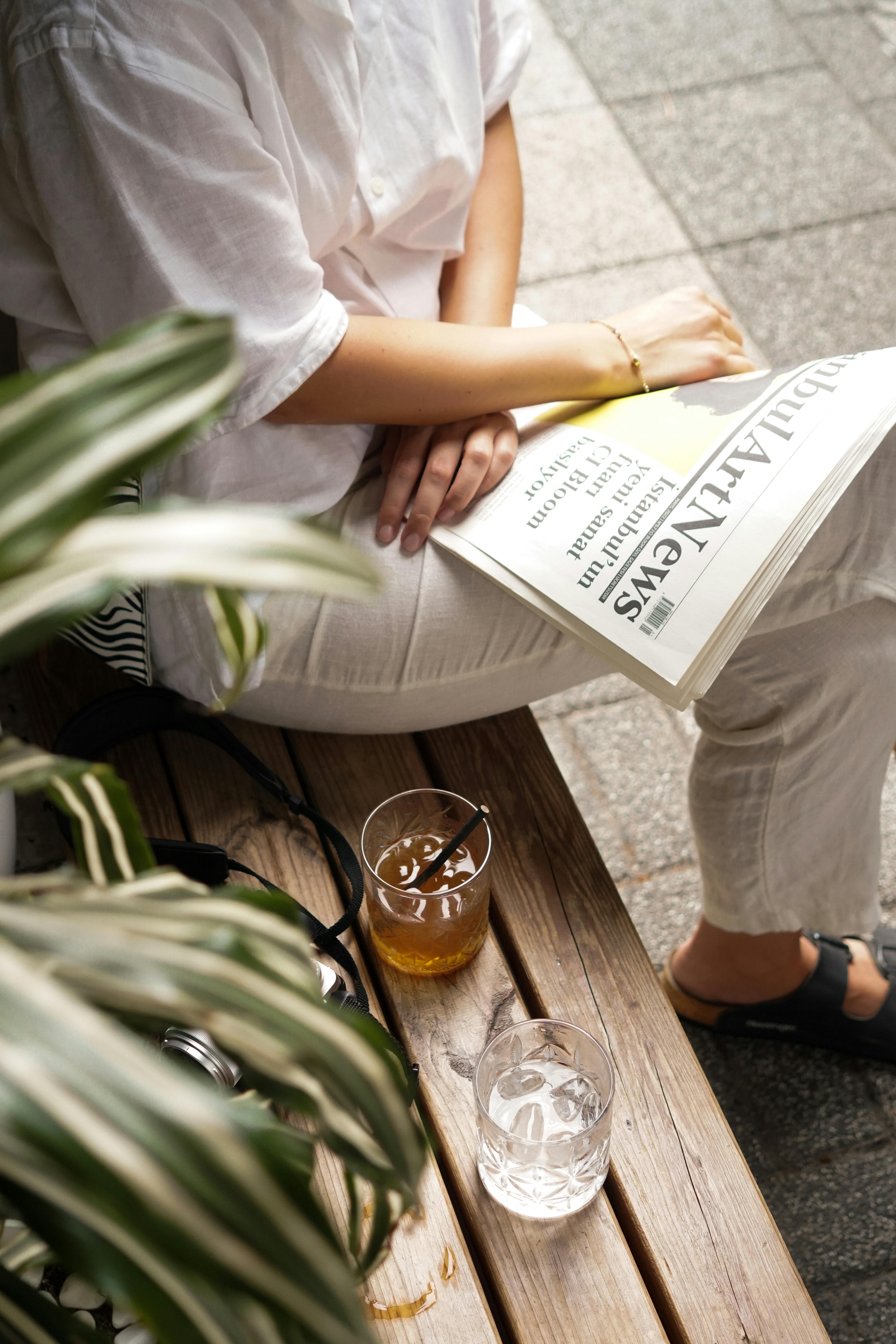 Woman sitting outdoors on a bench, reading a newspaper with a drink beside her.