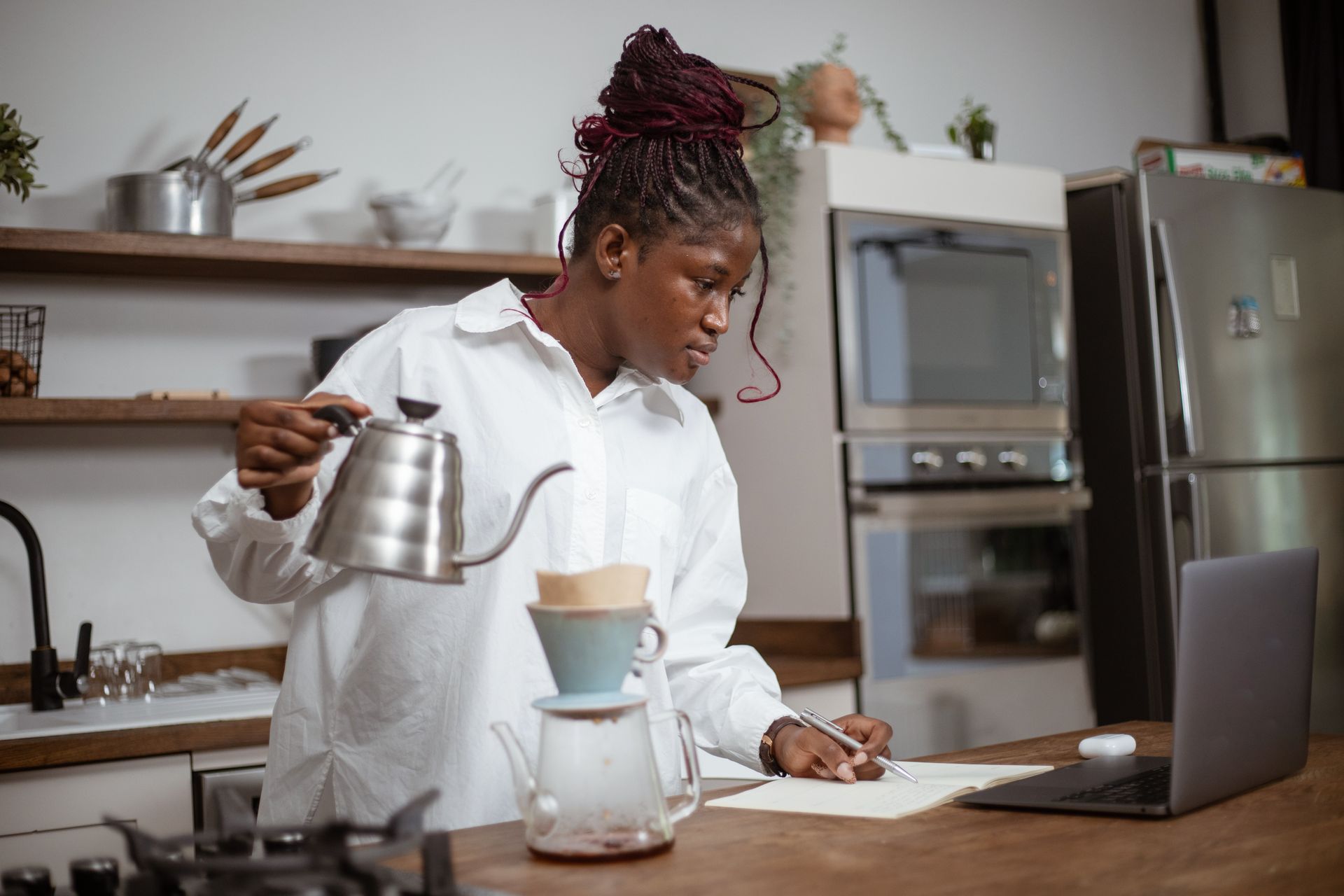 A woman pours coffee into a filter while looking at a laptop in a chic kitchen.