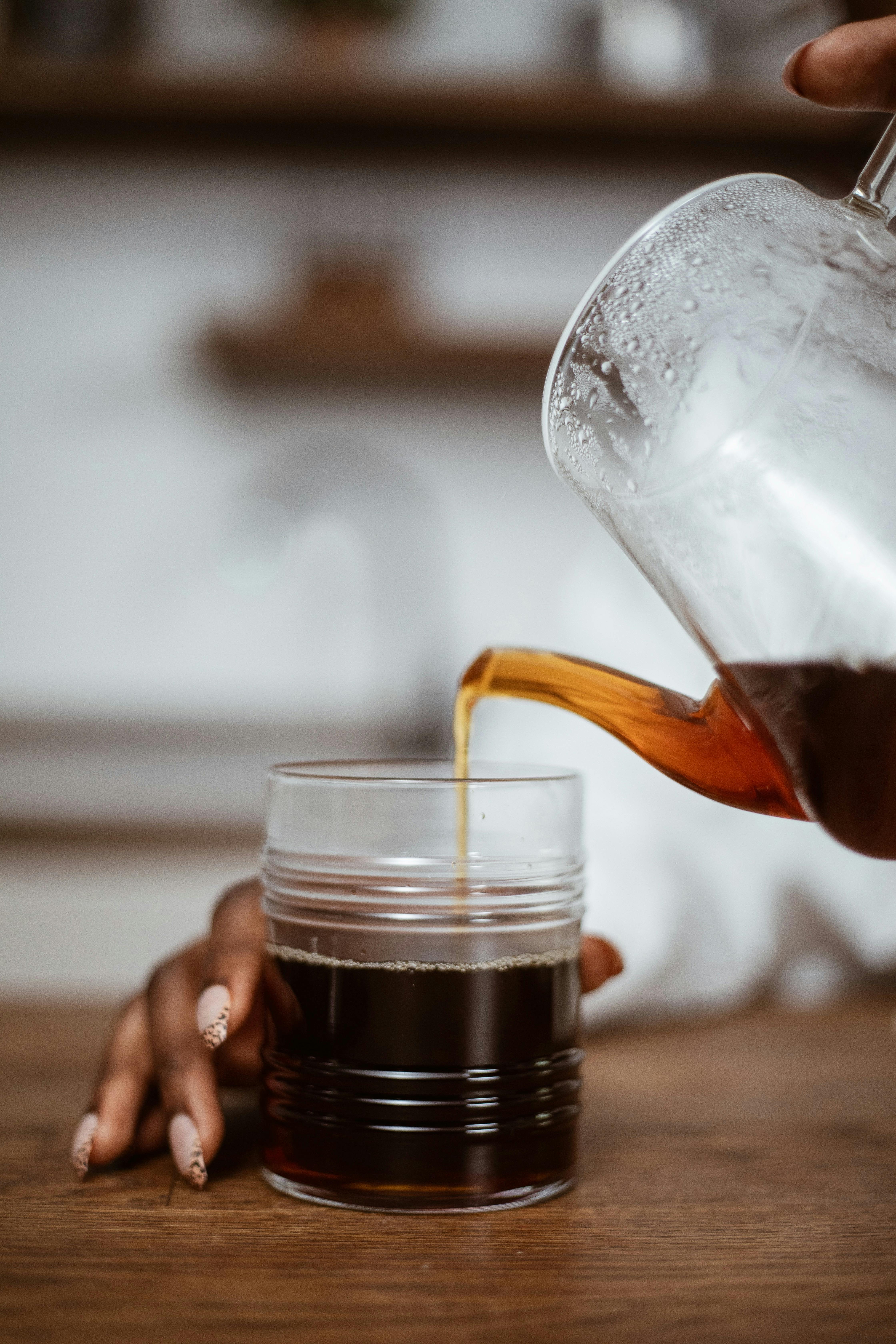 A person pours hot tea from a jug into a glass in a cozy kitchen setting.
