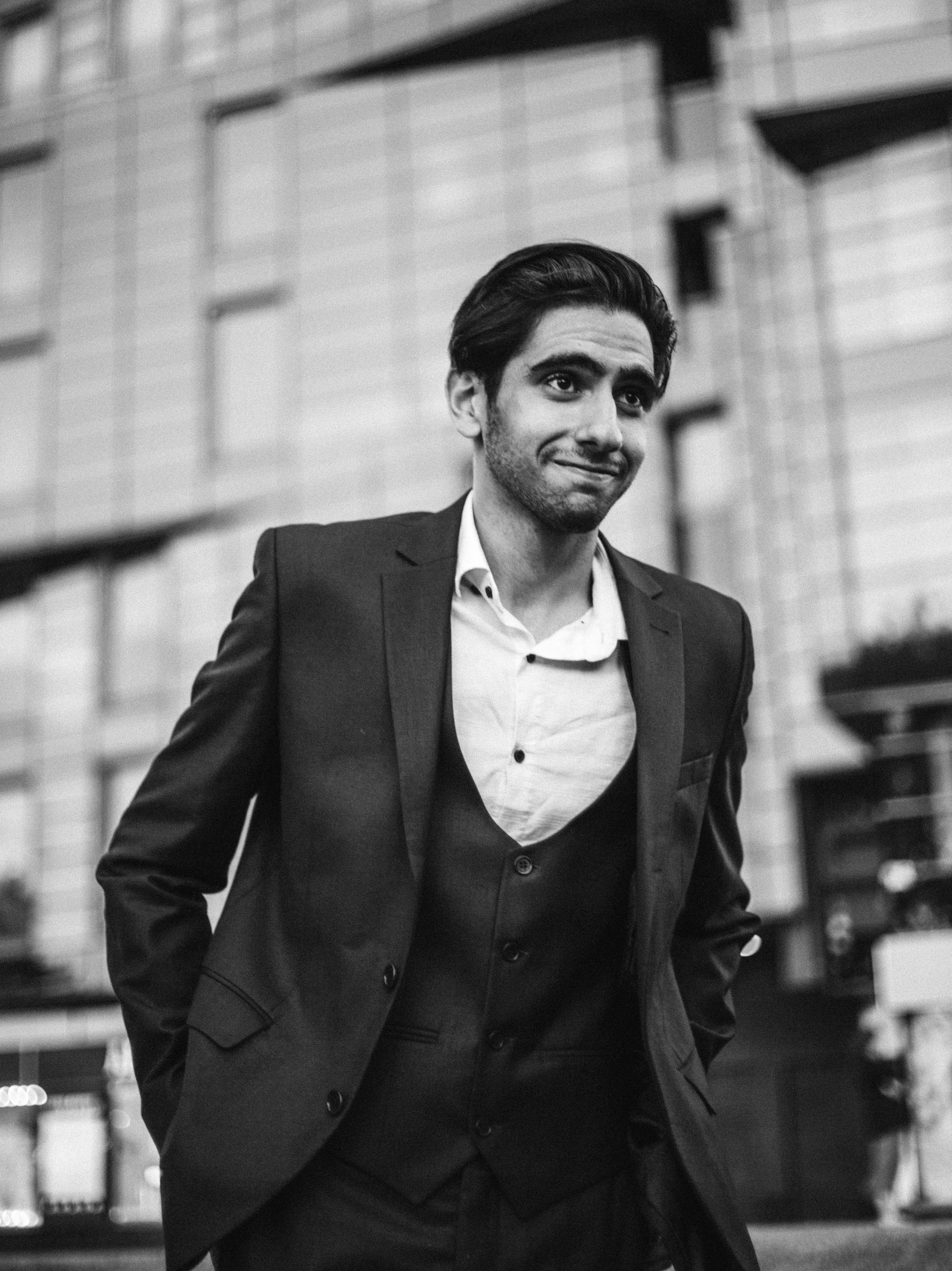 Black and white photo of an adult man in a suit smiling with hands in pockets.
