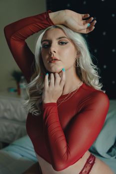 Blonde woman posing confidently in a red mesh top in a cozy bedroom setting.