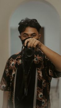 Portrait of an Asian man holding a camera indoors, featuring a stylish shirt and confident pose.