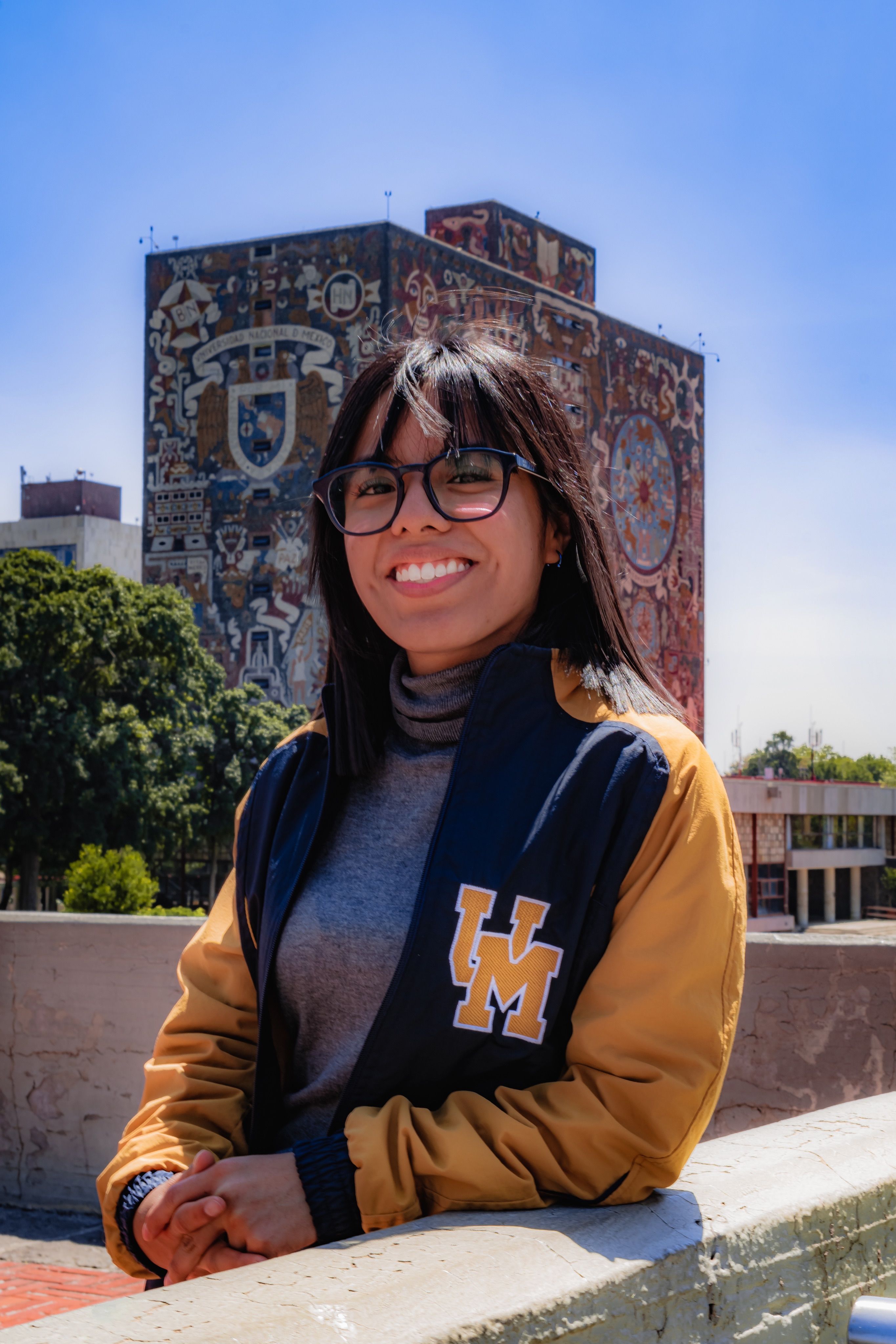 Young woman smiling in front of iconic UNAM mural in bright sunlight.