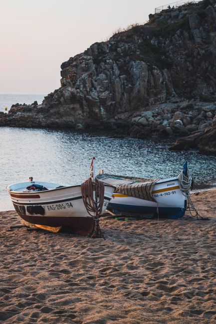 Two fishing boats rest on a sandy beach near cliffs, with a tranquil sea at sunset.