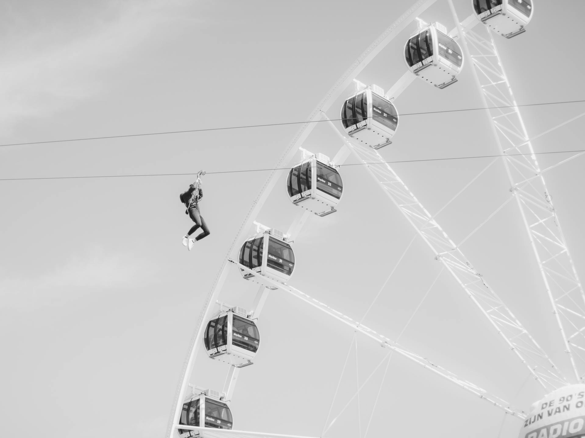 Thrilling black and white shot of a person zip lining past a Ferris wheel in a daring amusement park scene.