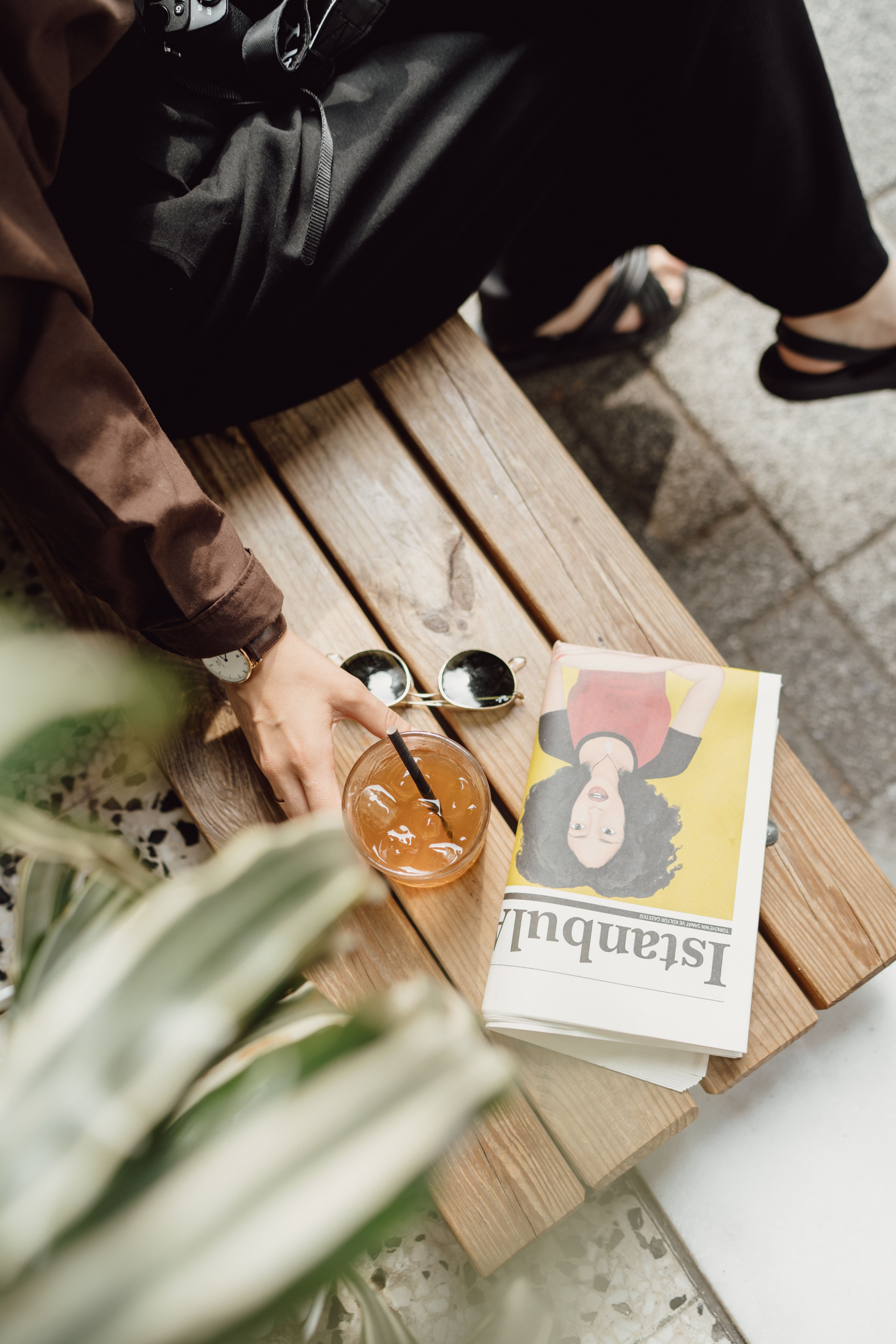 A person enjoys a drink while reading a newspaper on a wooden bench outdoors.