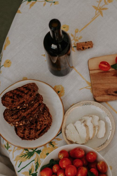 Free A cozy rustic dining setup featuring a wine bottle, bread, cheese, and cherry tomatoes on a floral tablecloth. Stock Photo