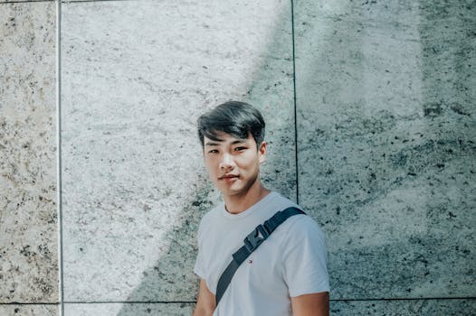 一位年轻亚洲男子的随意肖像，在户外阳光下倚靠着纹理石墙