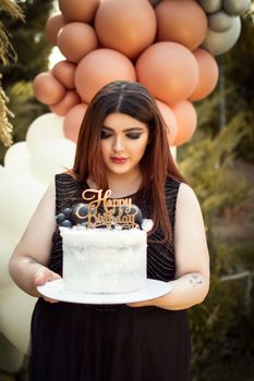 A woman holding a birthday cake with balloons in the background. Outdoor celebration in Tehran, Iran.