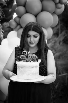 A woman holding a decorated cake at a birthday party with balloons in the background.