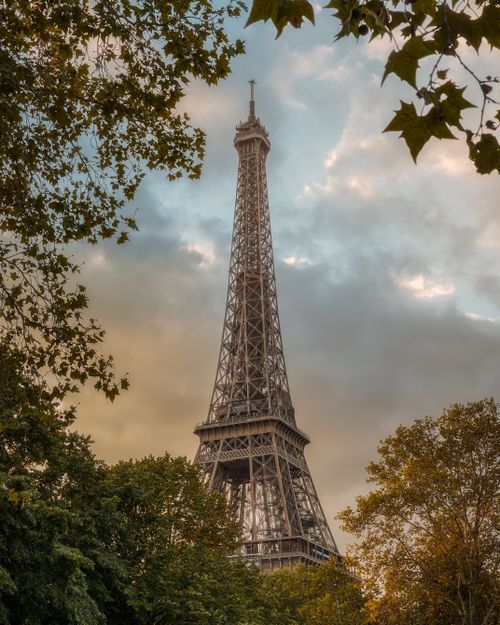 Free A stunning view of the Eiffel Tower framed by lush green trees under a blue and cloudy sky. Stock Photo