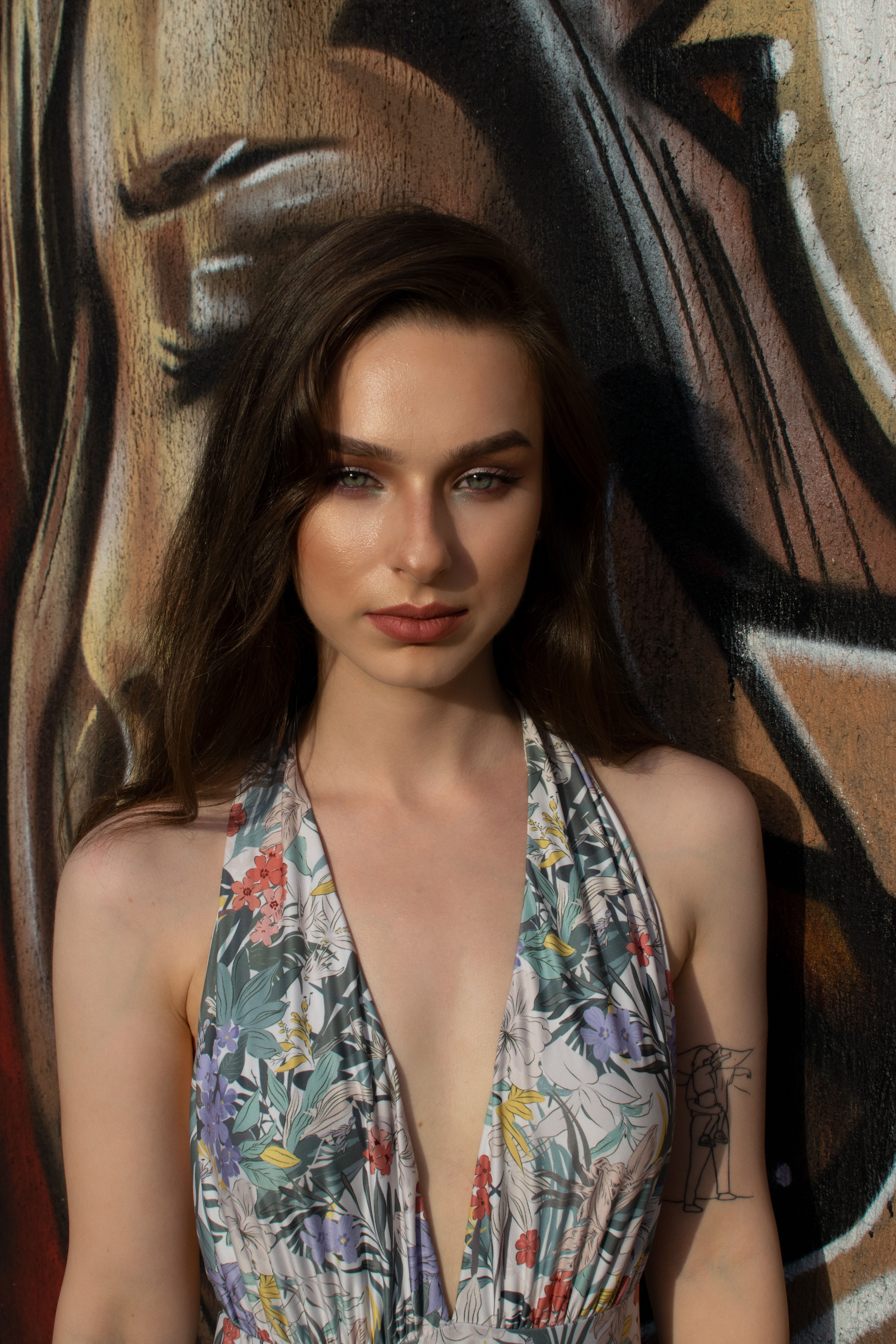 Stylish young woman in a floral blouse poses against a vibrant graffiti wall in the sun.