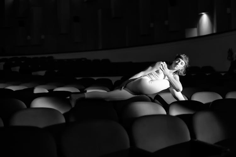 Black And White Photo Of Woman Stretching Across Empty Movie Theatre Seats